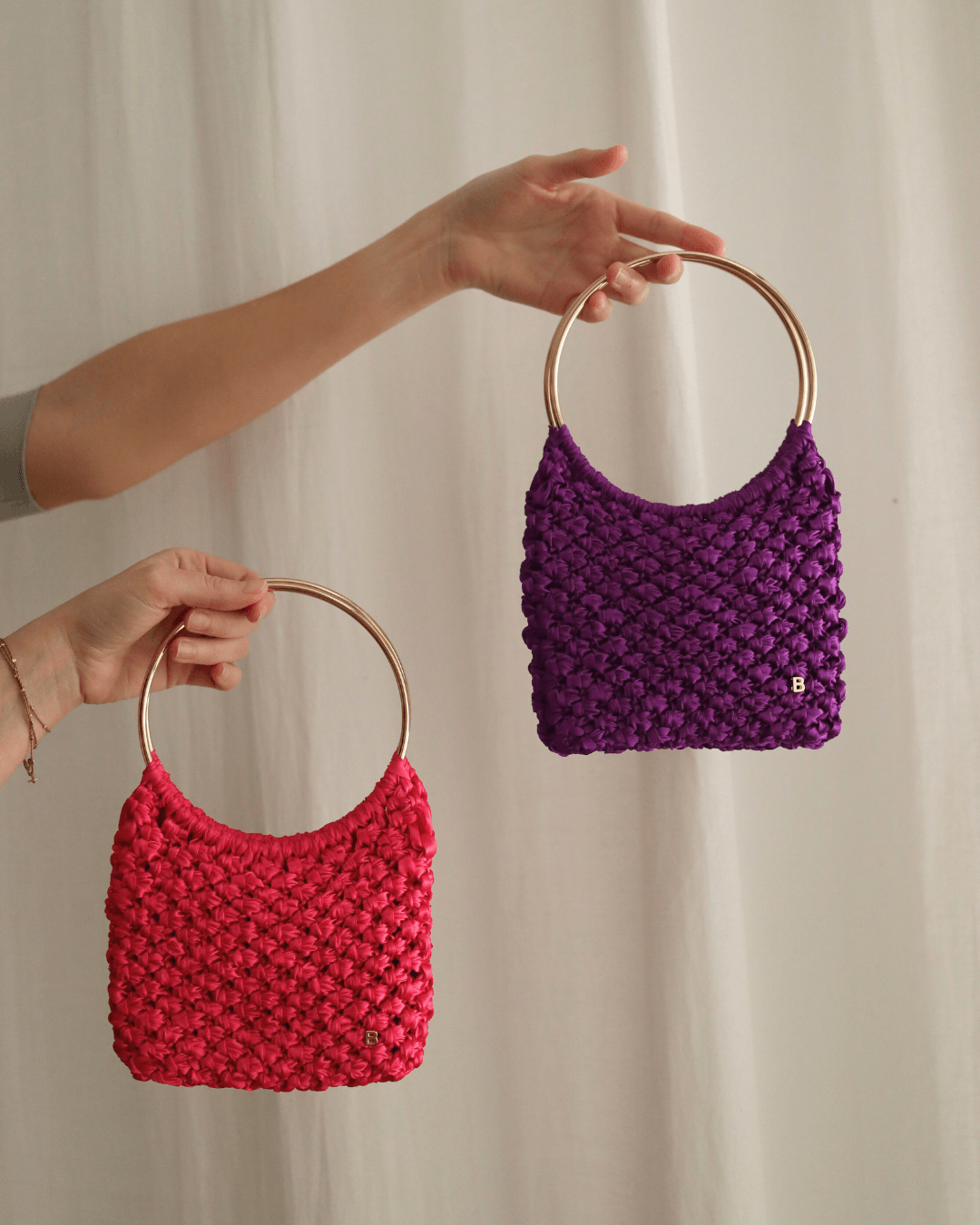 Roma Bag de satén fucsia y morado, hechos a mano con nudos de macramé, sujetados sobre fondo blanco.